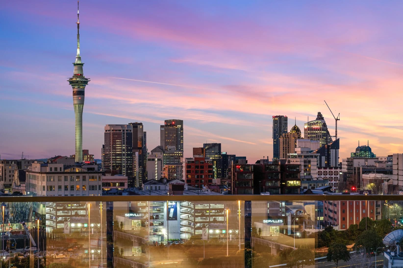 Auckland city skyline at sunset viewed from an Eden Terrace apartment balcony with glass railing reflections